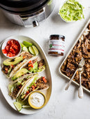 A platter with three tacos filled with shredded meat, lettuce, tomato, avocado, and a sweet-spicy kick from Terrapin Ridge Farms Jalapeno Hatch Chile Jam, served with lime wedges and sour cream. Nearby: extra jam, chopped tomatoes, lettuce, and shredded meat.