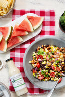A bowl of colorful bean and corn salad sits on a red checkered cloth next to a marble plate with sliced watermelon, a bottle of Terrapin Ridge Farms Cilantro Lime Ranch Salad Dressing, and a bowl of potato chips.