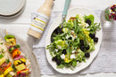 A fresh green salad with blackberries, avocado, feta, and nuts sits beside a bottle of Terrapin Ridge Farms Lemon Honey Vinaigrette, grilled veggie skewers with gluten free marinade, and a small vase of flowers on a white wooden table.