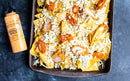 A tray of nachos with shredded cheese, blue cheese crumbles, and buffalo chicken sits next to a bottle of Terrapin Ridge Farms Buffalo Ranch Squeeze on a dark textured surface.