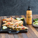 Grilled vegetable quesadillas on a cast iron tray, garnished with lime wedges. Nearby is a bottle of Terrapin Ridge Farms Buffalo Ranch Squeeze, plus bowls of chopped cilantro and avocado slices on a wooden table.