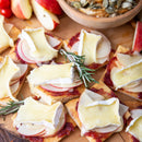 Sliced crackers with apple, brie, and Terrapin Ridge Farms Cranberry Relish With Grand Marnier™ are arranged on a wooden board and garnished with rosemary—ideal for holiday entertaining. Apple slices and seeds add festive flair to the scene.