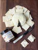 A ball of shredded cheese sits with square crackers on a wooden board, next to Terrapin Ridge Farms Apple and Horseradish Jam—a zesty touch for any gourmet spread—with two crackers topped with this creamy blend.