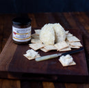 A wooden board displays a cheese ball, square crackers, and a jar of Terrapin Ridge Farms Apple and Horseradish Jam, with a knife holding cheese on a cracker.