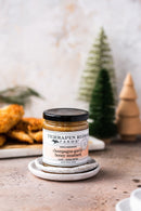 A jar of Terrapin Ridge Farms Champagne Garlic Honey Mustard sits on stacked ceramic plates, with blurred fried food and wooden trees in the background—ideal for charcuterie boards.
