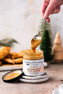 A hand scoops Terrapin Ridge Farms Champagne Garlic Honey Mustard with a spoon. This sweet, tangy condiment is perfect for charcuterie boards and fried foods, with a small Christmas tree blurred in the background.