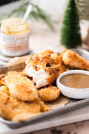 A tray of crispy breaded chicken tenders is served with Terrapin Ridge Farms' Champagne Garlic Honey Mustard. One tender is split open to reveal the juicy inside, with a jar of honey and a decorative pine tree in the background.