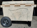 A large beige speckled cooler with a fish design and wheels, compatible with the Taiga Coolers Ice Chest Wheel Kit for 55quart and 88 quart coolers, sits on a concrete floor inside a building.
