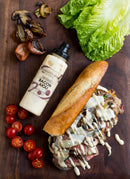 A baguette sandwich with grilled steak, mushrooms, and Terrapin Ridge Farms Bacon Aioli Squeeze sits on a board, surrounded by sliced grape tomatoes, grilled onions and mushrooms, lettuce leaves, and a bottle of Terrapin Ridge Farms Bacon Aioli.