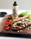 A steak sandwich on a wooden board, topped with Terrapin Ridge Farms Bacon Aioli Squeeze and served with cherry tomato halves and lettuce. A bottle of Terrapin Ridge Farms Bacon Aioli stands behind the sandwich.