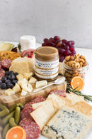 A charcuterie board with assorted cheeses, salami, crackers, pickles, nuts, grapes, and a jar of Terrapin Ridge Farms Pecan Honey Mustard in the center adds sweet nutty flavor. Sliced orange and rosemary sprigs garnish the board.