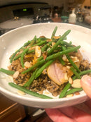 A bowl of rice and quinoa with green beans, sliced almonds, and seared pork, finished with a drizzle of Terrapin Ridge Farms Garlic Kraut Mustard, held up in a kitchen setting.
