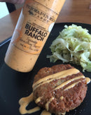 A bottle of Terrapin Ridge Farms Buffalo Ranch Squeeze is held over a plate with a fried patty topped with the creamy buffalo ranch sauce, served alongside cooked cabbage.