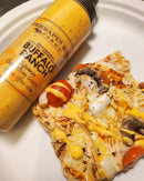 A bottle of Terrapin Ridge Farms Buffalo Ranch Squeeze sits beside a slice of pizza with onions, mushrooms, tomatoes, chicken, and a yellow sauce drizzle on a white plate.