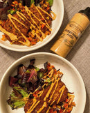 Two white bowls filled with leafy green salads, veggies, and crispy patties are drizzled with a creamy yellow sauce. Next to the bowls is a bottle of Terrapin Ridge Farms Everything Aioli Garnishing Squeeze on a beige fabric.