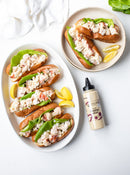 A platter and a plate of lobster rolls with lettuce in split-top buns, served with lemon wedges and Terrapin Ridge Farms Truffle Aioli Squeeze as a gourmet finishing sauce on a white surface.