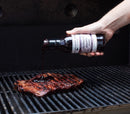 A hand pours Terrapin Ridge Farms Blackberry Chipotle Sauce from a bottle onto a steak sizzling on the grill, with the flavorful sauce spreading over the meat.