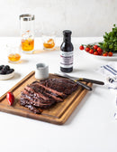 Sliced grilled steak on a wooden board with knife, fork, and sauce jar. Terrapin Ridge Farms Blackberry Chipotle Sauce, glasses of whiskey, blackberries, cherry tomatoes, and greens are arranged on a white table.