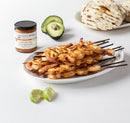 A plate of grilled shrimp skewers sits on a white table with lime wedges, an avocado half, Terrapin Ridge Farms Nashville Hot Mustard, and a stack of grilled flatbreads in the background.