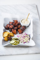 A tray lined with parchment holds glazed meat, roasted potatoes, coleslaw, pickles, and a bowl of Terrapin Ridge Farms Truffle Aioli Squeeze by Terrapin Ridge Farms with a gold spoon, all arranged on a marble surface.