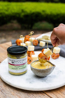 A hand dips a sausage and cheese skewer into a bowl of Terrapin Ridge Raspberry Wasabi Mustard, highlighting this sweet heat condiment by Terrapin Ridge Farms. A nearby jar and arranged skewers rest on a marble board.