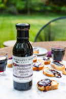 A bottle of Terrapin Ridge Farms Blackberry Chipotle Sauce sits on a table outdoors with sliced bread topped with the sauce and two glasses of red wine, framed by lush greenery.