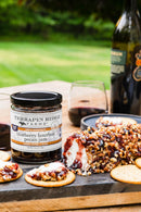 Terrapin Ridge Farms Blueberry Bourbon Pecan Jam is paired with goat cheese coated in jam and pecans, served with crackers on a wooden board—ideal for charcuterie. Red wine bottles and glasses are seen in the background.