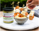 A hand dips a skewer of sausage and cheese into creamy Terrapin Ridge Farms Raspberry Wasabi Mustard. Nearby is a jar of the mustard, with more skewers arranged on a white marble platter in the background.