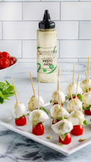 Terrapin Ridge Farms Pesto Aioli Squeeze 12.5 oz stands behind skewers of cherry tomatoes, mozzarella, and basil drizzled with the sauce. A bowl of tomatoes and basil sits on a white countertop with a tiled background.