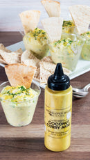 A bottle of Terrapin Ridge Farms Coconut Curry Aioli is displayed with small cups of the creamy sauce, garnished with chopped herbs and pita chips, all arranged on a wooden surface with a white plate in the background.