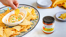 A hand dips a tortilla chip into a creamy dip with an orange swirl, surrounded by more chips. Terrapin Ridge Farms Mango Habanero Jam from Terrapin Ridge Farms sits nearby—an ideal sweet and spicy addition for appetizers. Plates and napkin are in the background.