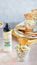 A clear plastic cup of pasta salad and pita chips sits on a table next to a Terrapin Ridge Farms Avo Aioli Roasted Garlic Squeeze bottle, with more cups, pita, and a yellow checkered napkin in the background for a bright, inviting setting.