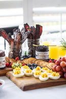 A breakfast spread features deviled eggs, grapes, berries, kiwi, bacon in jars, pancakes, Terrapin Ridge Farms Blueberry Bourbon Pecan Jam, and orange juice served on a wooden board.