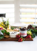 A wooden cheese board with brie topped with Terrapin Ridge Farms Strawberry & Fig Jam, fresh strawberries, cookies, and holly, set on a white tablecloth with festive greenery and pinecones in the background.