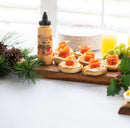 A wooden serving board with crackers topped with cream and smoked salmon, Terrapin Ridge Farms Sriracha Horseradish Garnishing Squeeze Sauce for garnishing, green grapes, and festive greenery on a white tablecloth. Plates and orange juice are in the background.
