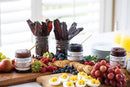 A breakfast spread with deviled eggs, berries, grapes, and pancakes is topped with Terrapin Ridge Farms Raspberry Amaretto Jam. Jars of bacon strips, juice, and fruit spread are on the side; plates are stacked in the background.