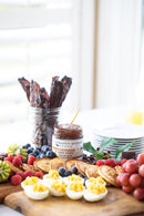 A breakfast charcuterie board highlights deviled eggs, pancakes, fruit, and Terrapin Ridge Farms Apple Maple Bacon Jam. In the background are strips of bacon, savory jam, stacked plates, and orange juice.