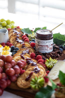 A brunch platter features pancakes topped with Terrapin Ridge Farms Raspberry Amaretto Jam, fresh grapes, berries, deviled eggs, pecans, and a jar of Terrapin Ridge Farms jam with a spoon on a wooden board—ideal for gourmet desserts.