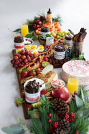 A festive cheese board with fruit, crackers, deviled eggs, bacon, drinks, and Terrapin Ridge Farms Strawberry & Fig Jam is surrounded by pinecones, greenery, and holiday decor on a white surface.