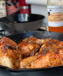 Chicken pieces cooked in a cast iron skillet and glazed with Terrapin Ridge Farms Hot Pepper Peach Bourbon Sauce. A bottle of the sauce and a skillet are visible in the background.