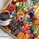 A wooden platter features assorted cheeses, meats arranged as roses, fresh fruits, figs, cookies, nuts, edible flowers, herbs, and a jar of Terrapin Ridge Farms Bacon Pepper Dip for a colorful appetizer display.