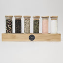 The Spice Wheel Salt & Pepper Collection features six glass jars with wooden lids, filled with peppercorn medley and pink Himalayan sea salt, elegantly arranged in a bamboo box holder against a white background.