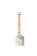 The Smokin Brothers SB Burger Flipper by Smokin Brothers is a versatile grilling tool featuring a sturdy metal spatula with a wooden handle, a convenient bottle opener at the top, and SB cutouts on the flat metal surface—perfect for flipping burgers or using as a griddle accessory.