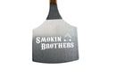 Introducing the Smokin Brothers Butt Lifter—a heavy-duty metal pizza peel with a sturdy wooden handle. The blade features “Smokin Brothers” and a small flame graphic, both printed in white. Perfect not only for pizzas but also as a BBQ meat lifter or pork butt lifter, this versatile tool is an essential addition to any grilling toolkit.