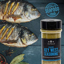 A bottle of The Spice Lab Taste of America Seasoning Collection – Key West Seasoning sits beside two grilled fish on a dark surface, surrounded by herbs and spices. Its blue label reads “Perfect for Seafood Cooking.” This American-inspired blend from The Spice Lab is ideal for grilling and enhances every bite.