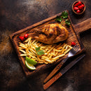 Grilled chicken and French fries served on a wooden board with lime wedges, parsley, and small red peppers sprinkled with The Spice Lab Birdseye Chili Pepper Powder by The Spice Lab, accompanied by a fork and knife, all set on a dark rustic surface.