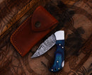 The Texan Knives Personalized Damascus Steel Pocket Knife, featuring a patterned blade and blue handle, is shown beside a brown leather case on a carved wooden surface—ideal as a custom Valentine’s Day gift for your husband.