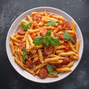 A white plate filled with penne pasta, sliced sausage, cherry tomatoes, arugula leaves, grated cheese, and a fresh basil garnish—finished with a sprinkle of The Spice Lab Organic Crushed Red Pepper Flakes from The Spice Lab for extra zest—set on a dark textured surface.