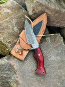 The Texan Knives Hunting Skinning Knife features a 10" Damascus steel blade and polished pakkawood handle, resting on its brown leather sheath atop rugged stone outdoors.