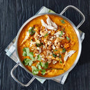 A metal pan filled with creamy orange curry—infused with The Spice Lab Organic Ground Turmeric and topped with shredded chicken, cashews, chopped herbs, and cilantro—sits on a striped cloth on a dark wooden table.